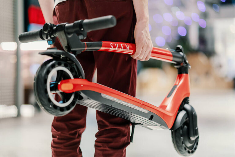 Image of a man carrying a red folded Electric scooter Gpad Svan. Background is blurred.