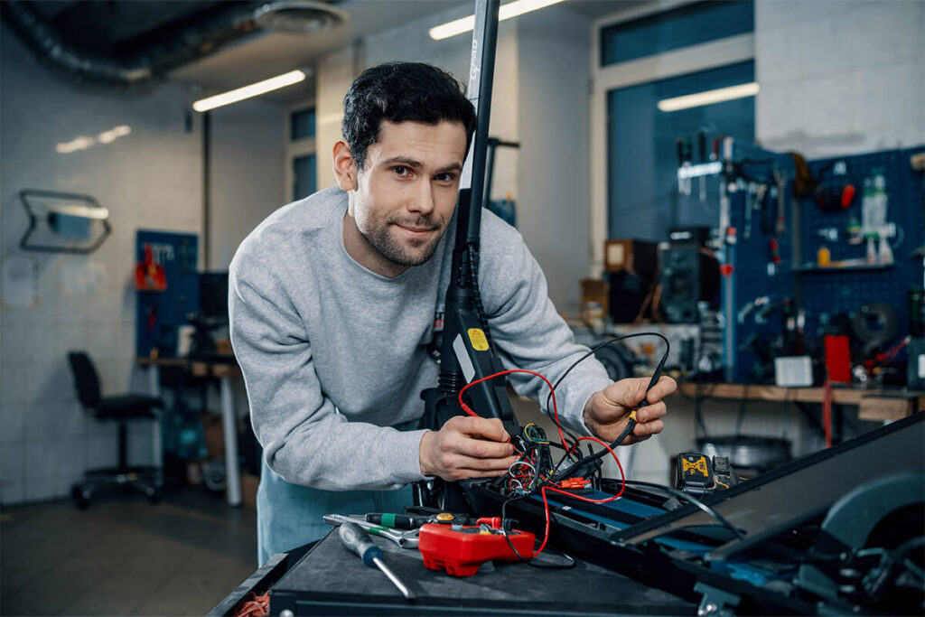 Technician repairing electric scooter wiring, surrounded by tools and electronic components in a detailed service workspace.