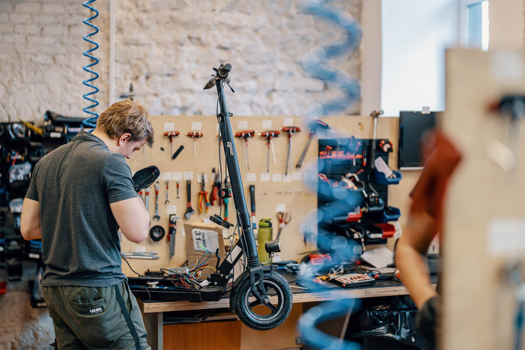Technician working on disassembled electric scooter at a service bench with tools hanging on the wall in the background.