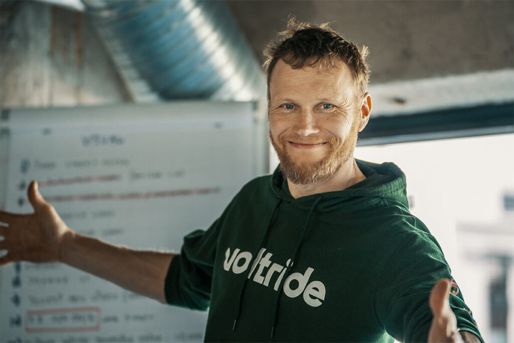 Voltride founder Mart Einpalu smiling and gesturing warmly in front of a whiteboard, wearing a branded green hoodie inside a modern office space.