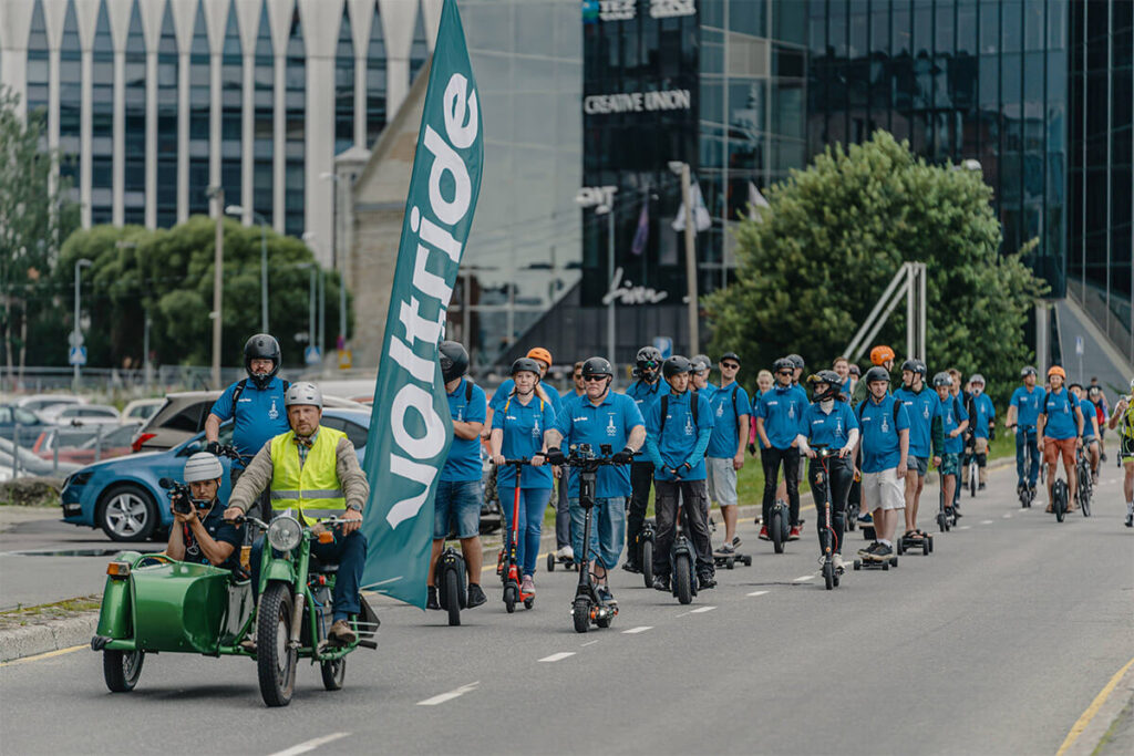 Large group of riders in blue Voltride shirts participating in a micromobility parade through a city street, led by a motorcycle with a sidecar and Voltride flag.