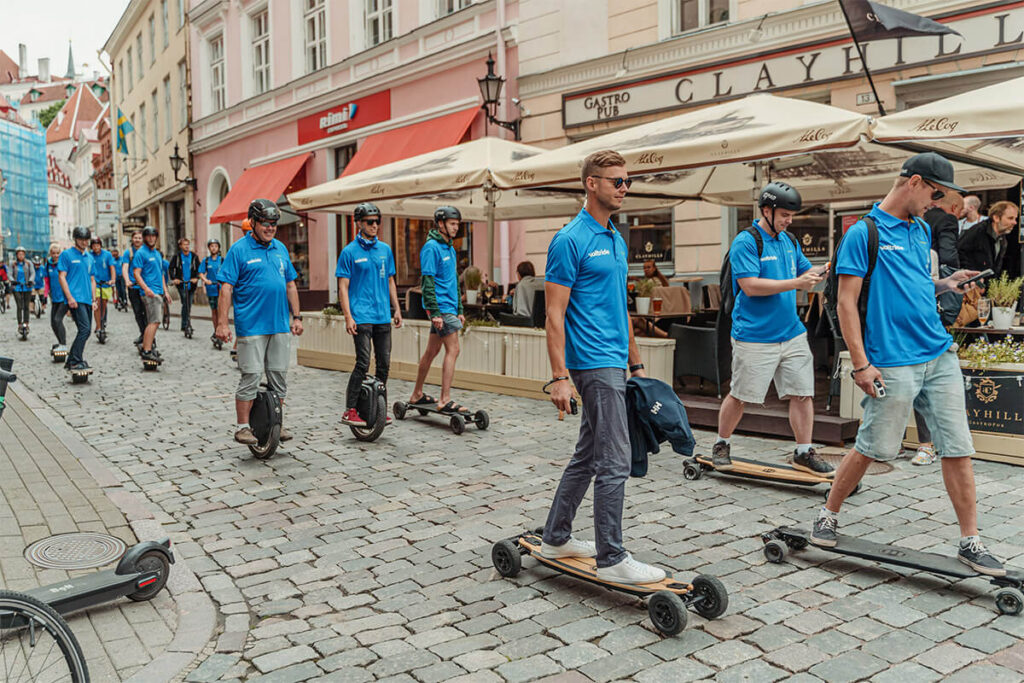 Voltride enthusiasts riding electric unicycles, skateboards, and scooters through the cobblestone streets of Tallinn Old Town, all wearing matching blue Voltride shirts and helmets.