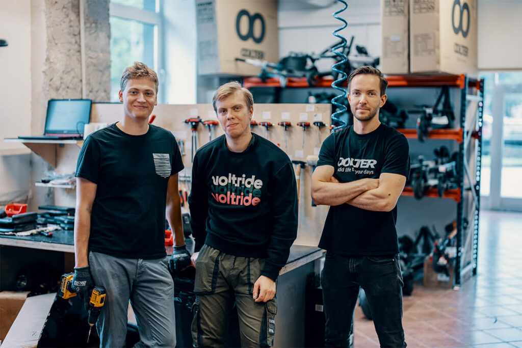 Three Voltride service technicians standing in a repair workshop, posing confidently with tools and branded shirts.