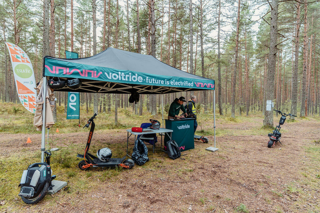 Voltride event setup in a forest with branded tent, electric scooters, and safety gear, serving as a checkpoint for an off-road micromobility competition.