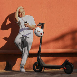 Smiling man leaning against an orange wall with Xiaomi electric scooter and helmet beside him