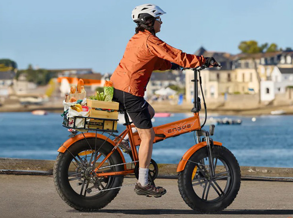 A person wearing a helmet rides an orange Engwe EP-2 Boost fat-tire electric bicycle near the waterfront, carrying a crate of groceries on the rear rack demonstrating the bike's cargo capacity.