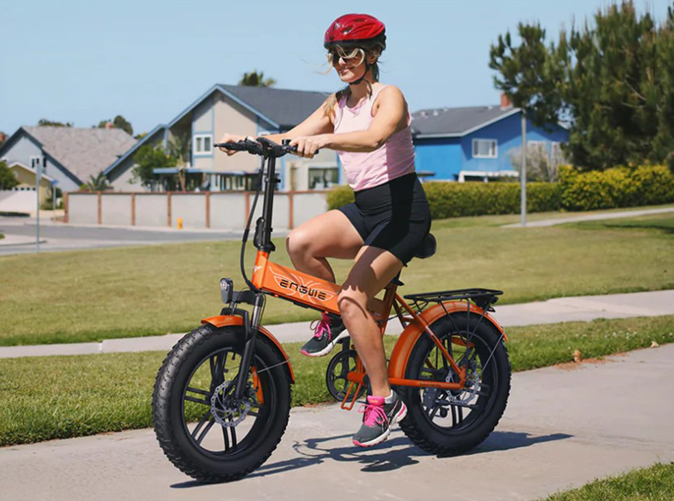 A woman wearing a helmet and sporty clothing rides an orange Engwe EP-2 Boost fat-tire electric bicycle on a sunny suburban path.