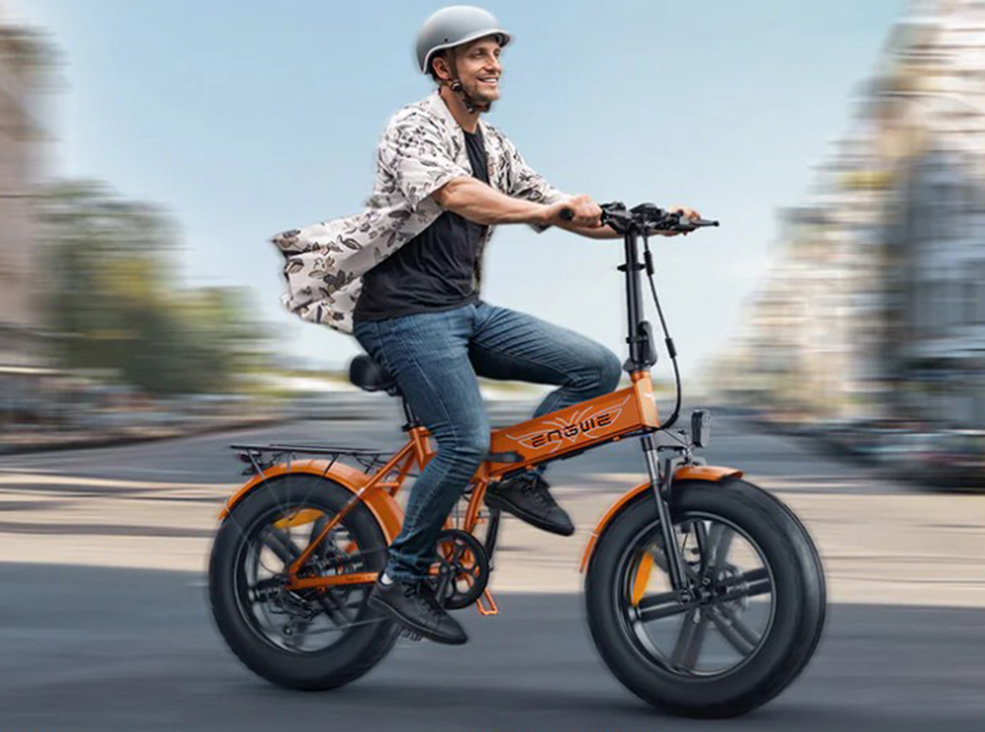 A smiling man wearing a helmet rides an orange Engwe EP-2 Boost E fat-tire electric bicycle through a city street, with the background blurred to show motion.