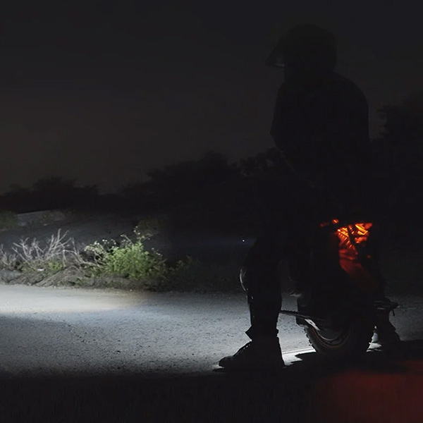 A rider on the Inmotion V14 electric unicycle travels on a dark road at night, illuminated by a bright forward headlight and a glowing rear light.