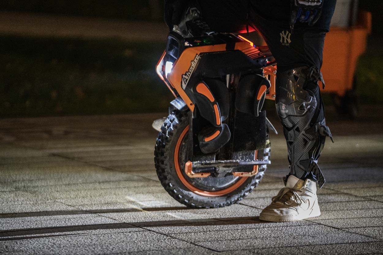 A rider wearing protective gear standng on the Inmotion V14 electric unicycle at night, with the headlight illuminating the ground and the knobby tire clearly visible.