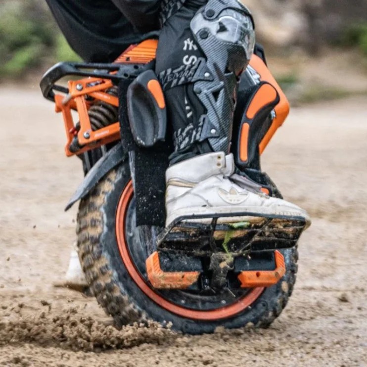 Close-up of a rider wearing protective gear and boots maneuvering the Inmotion V14 electric unicycle on a dirt track, showing the rugged tire and suspension design.