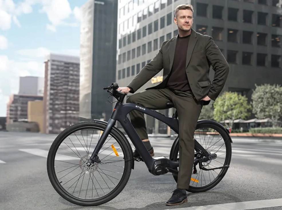 A man in a dark business suit sits on a black Engwe Mapfour N1 Pro electric bicycle in the middle of a city street, surrounded by tall modern buildings.