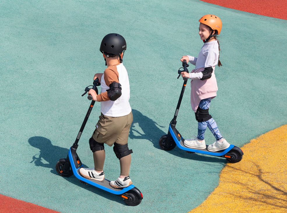 Two kids wearing helmets and protective gear riding the Segway-Ninebot C2 Lite Kids electric scooters.