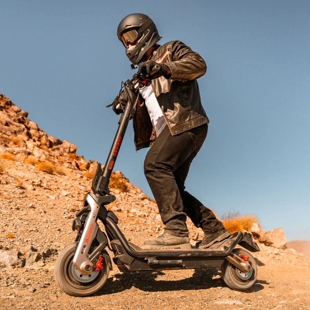 Rider wearing a helmet and leather jacket rides a Segway-Ninebot GT3 Pro electric scooter up a rocky hill, showcasing its powerful off-road performance.