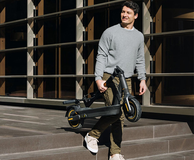 Person carrying a folded Segway-Ninebot MAX G2 E electric scooter down a few outdoor steps in a city area.