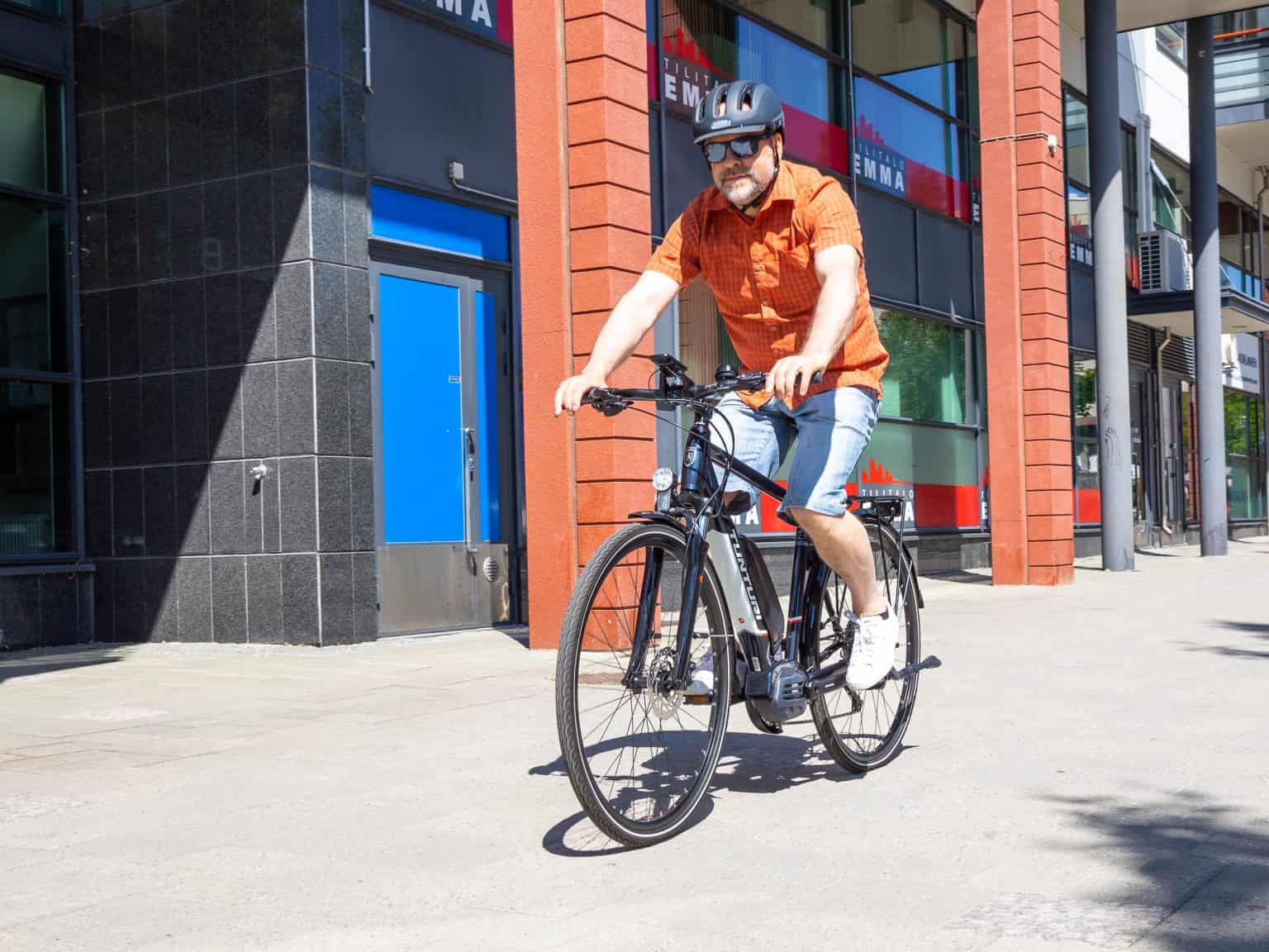 A man wearing a helmet rides the Tunturi eMotion e-bike through an urban area with modern buildings and bright sunlight.
