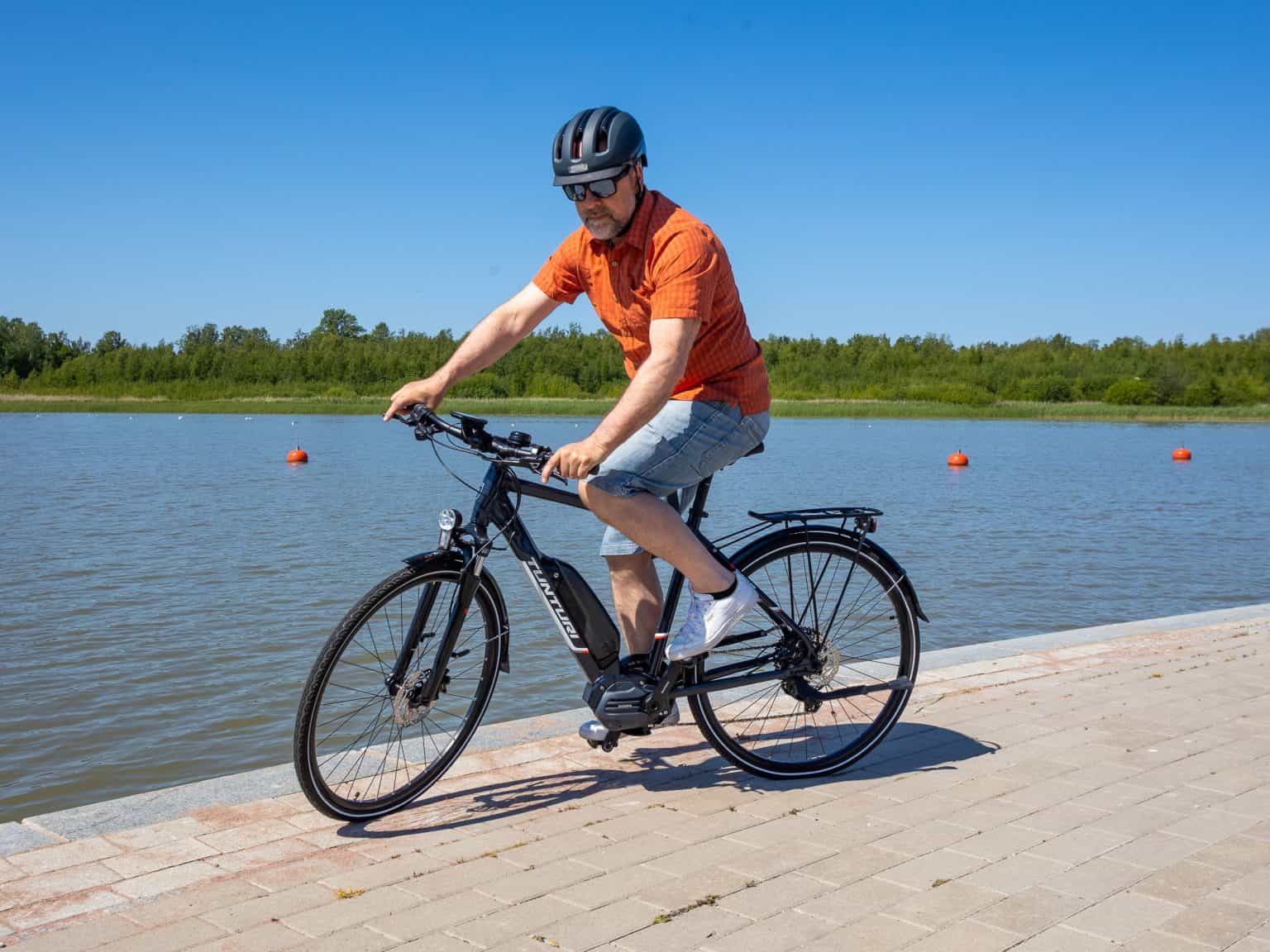 A man wearing a helmet rides a Tunturi eMotion electric bike along a paved path beside a calm waterfront on a clear, sunny day.