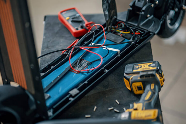 An open electric scooter deck showing the blue battery pack, wiring, and tools including a multimeter and a cordless drill used for diagnostics and repair.