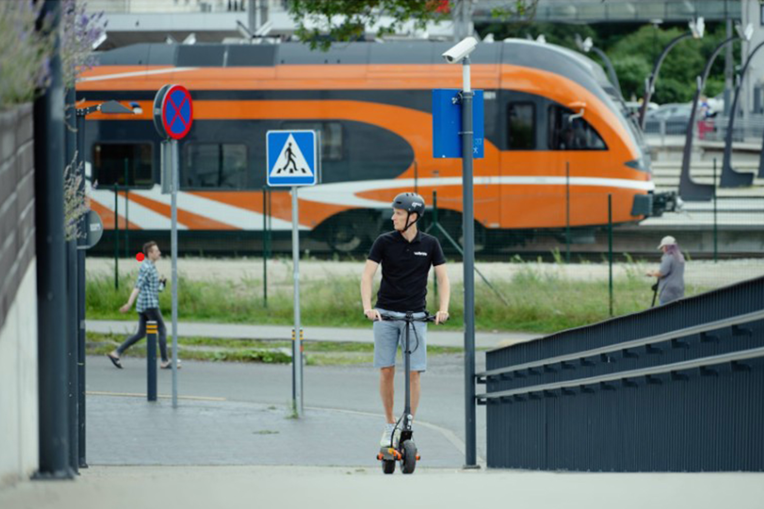 A man wearing a helmet rides an electric scooter along a city sidewalk near a railway station, with an orange commuter train passing in the background.
