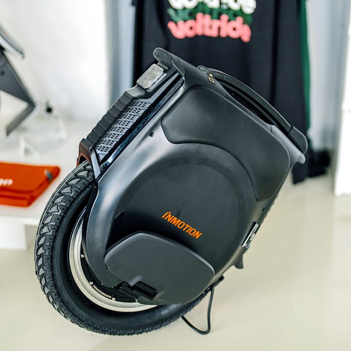 Inmotion V12 electric unicycle standing indoors, showing its black body, treaded wheel, side padding, and top handle, with Voltride merchandise blurred in the background.