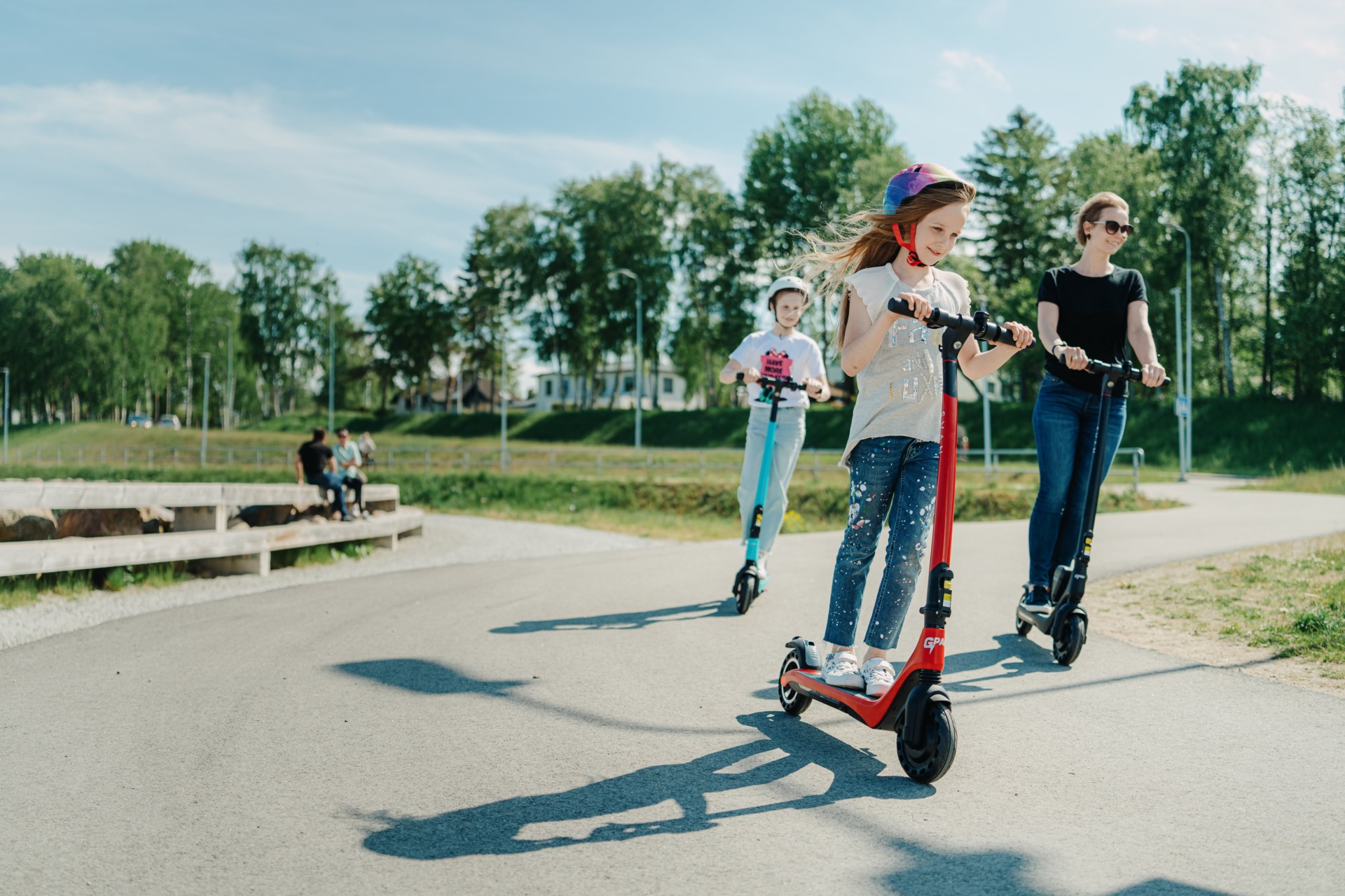 A child rides a red GPad Svan electric scooter on a sunny path, followed by another child and an adult on scooters in a park.