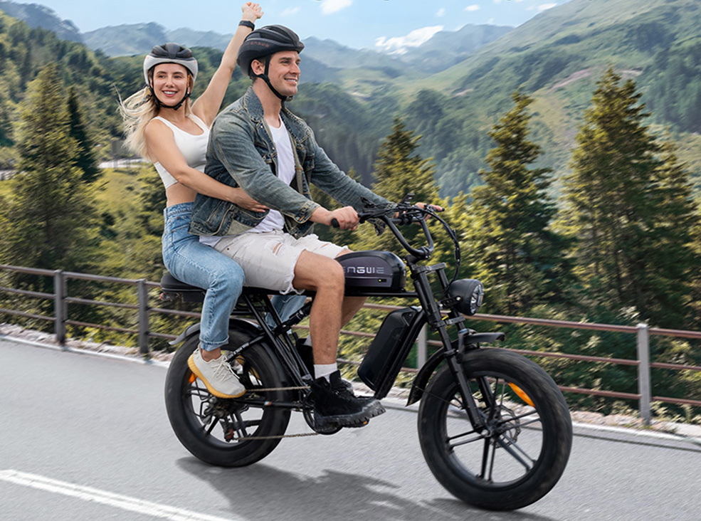 Two people riding the ENGWE M1 black fat-tire electric bike on a mountain road, both wearing helmets, with scenic forested hills in the background.