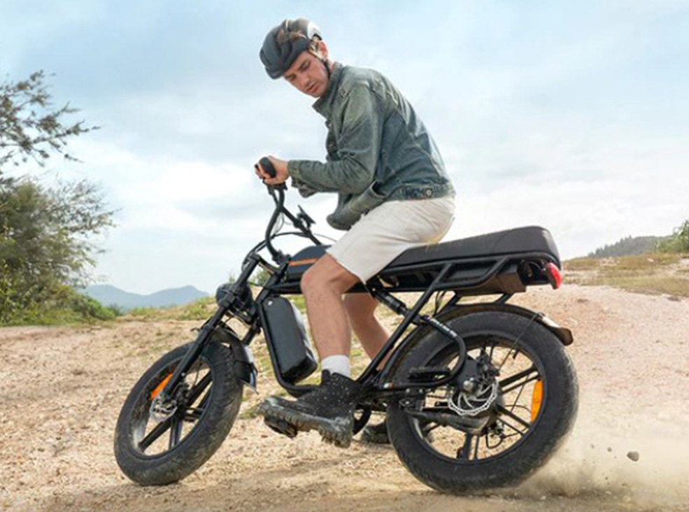 Man riding the ENGWE M1 black fat-tire electric bike on a dirt road, kicking up dust, wearing a helmet, green jacket, and light shorts in an outdoor landscape.