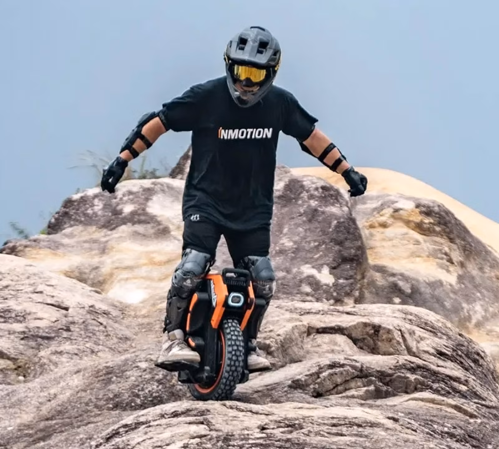 A person wearing a helmet, pads, and gloves riding an electric unicycle over large rocks outdoors